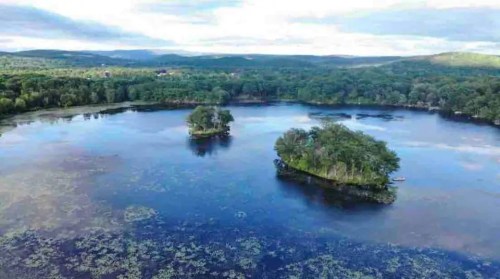 Aerial view of a serene lake with two small islands surrounded by lush greenery and distant hills.