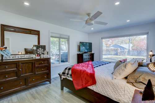 A bright bedroom featuring a bed with decorative pillows, a dresser, a TV, and large windows overlooking a backyard.