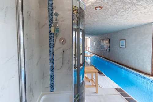 A modern shower stall beside a long indoor swimming pool, with light-colored walls and a wooden table nearby.