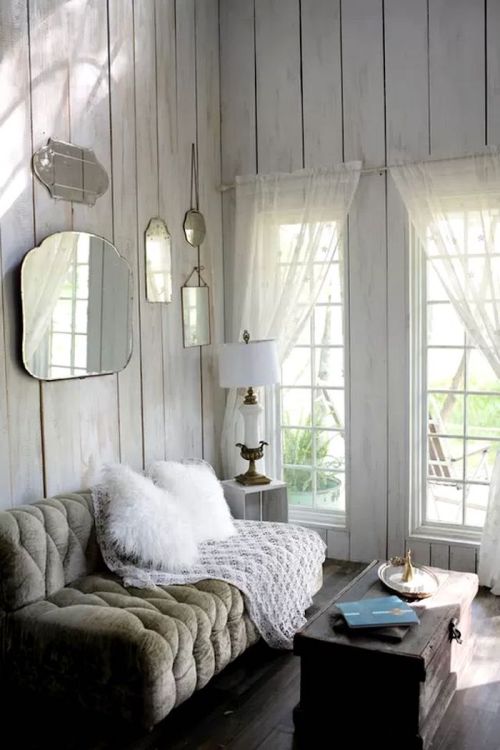 Cozy living room with a plush sofa, white throw, vintage mirrors, and large windows letting in natural light.