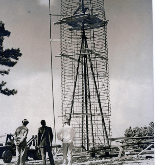 Three men observe a tall, partially constructed tower surrounded by trees and equipment in a vintage black-and-white photo.