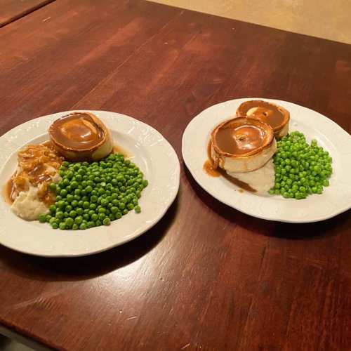 Two plates of food featuring green peas, mashed potatoes, and round meatloaf topped with gravy, on a wooden table.