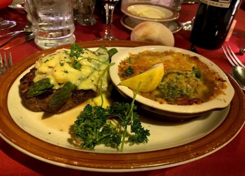 A plate featuring a steak topped with asparagus and hollandaise sauce, alongside a baked dish with cheese and herbs.