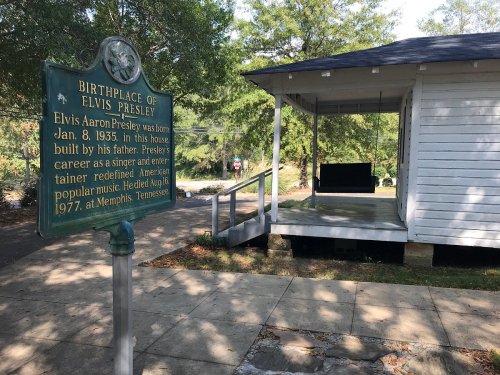 Sign marking the birthplace of Elvis Presley, with a small white house in the background, surrounded by trees.