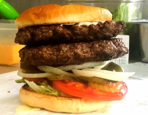 A towering burger with two beef patties, lettuce, tomato, pickles, onions, and mayonnaise on a toasted bun.
