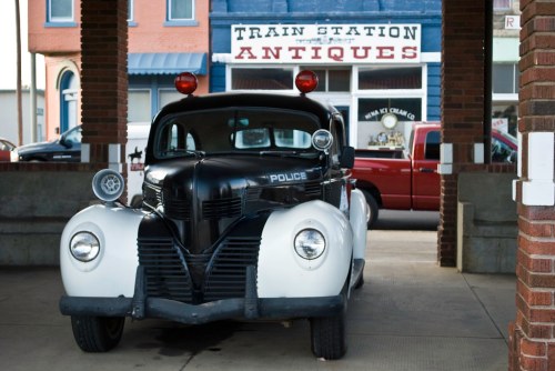 A vintage police car parked under a train station, with an antique shop in the background.