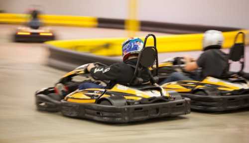 Two go-karts race around a track, with one driver in a blue helmet and the other in a silver helmet.