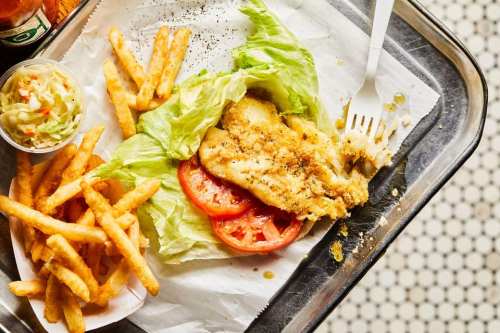A plate with a fried fish sandwich on lettuce and tomato, served with fries and a side of coleslaw.