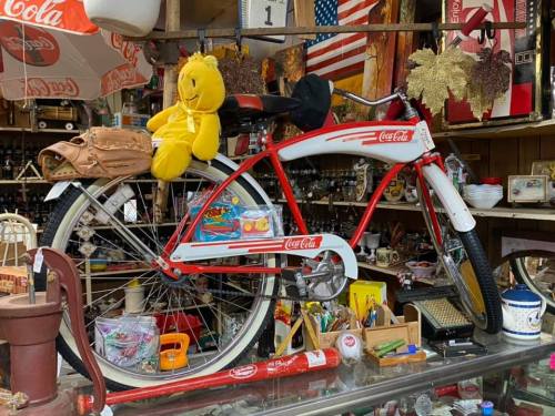 A vintage Coca-Cola bicycle displayed in a cluttered shop, adorned with a yellow stuffed animal and various collectibles.