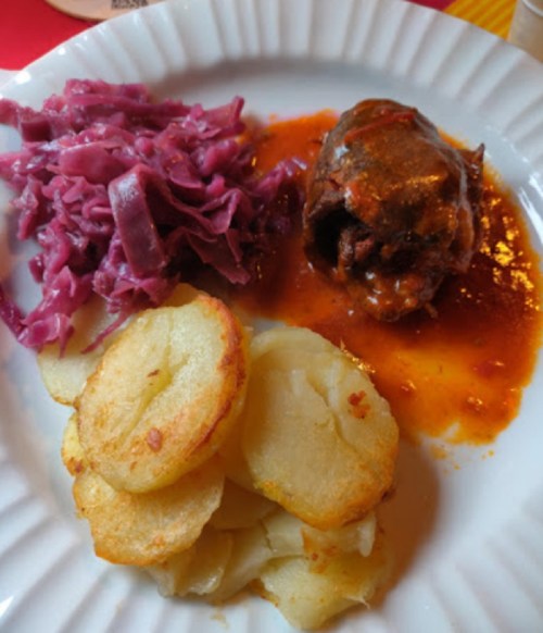 A plate featuring rolled meat in sauce, red cabbage, and two types of sliced, cooked potatoes.