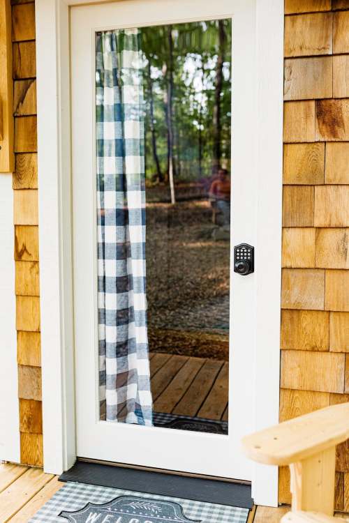 A white door with a glass panel, featuring a checkered curtain and a keypad lock, set against wooden siding.
