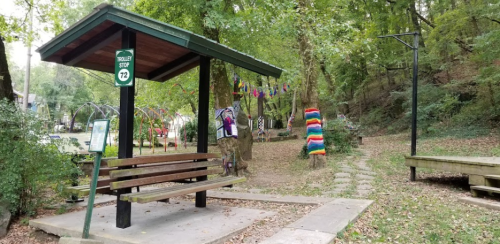A bus stop shelter with a bench surrounded by trees, colorful decorations, and a pathway in a serene park setting.