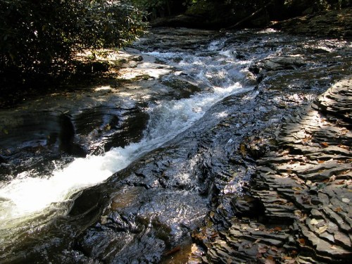 A serene river flows over rocky terrain, surrounded by lush greenery in a peaceful natural setting.