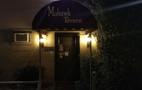 Entrance of Mohawk Tavern with a red awning, illuminated by warm lights, surrounded by greenery at night.