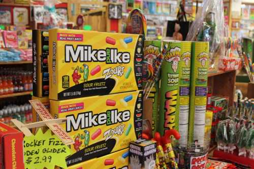 Colorful candy boxes of Mike and Ike Zours stacked in a vibrant store filled with various sweets and snacks.