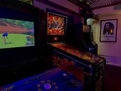 A dimly lit arcade featuring a golf video game, pinball machines, and a poster of Bob Marley on the wall.