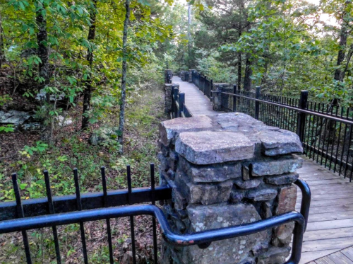 A winding wooden path surrounded by trees, with stone and metal railings along the sides.