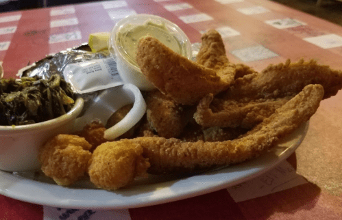 A plate of fried fish fillets, hushpuppies, collard greens, and a side of dipping sauce on a checkered tablecloth.