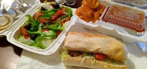 A takeout meal featuring a sub sandwich, salad with peppers and olives, and fried dough balls in a container.