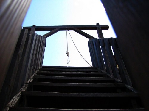 View from below a wooden staircase leading to a gallows with a noose hanging above against a bright sky.