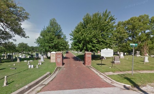 A tree-lined road leads into a cemetery, with gravestones visible on either side and a sign at the entrance.