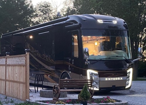 A sleek black motorhome parked near a wooden fence, surrounded by greenery and flowers.