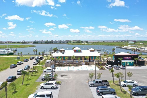 A colorful waterfront building with palm trees and parked cars, overlooking a calm bay under a blue sky with clouds.