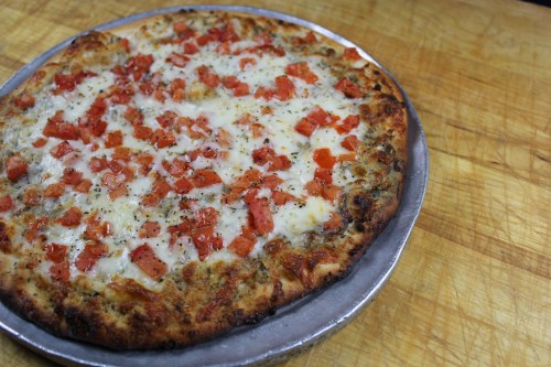 A freshly baked pizza topped with melted cheese and diced tomatoes on a silver tray, resting on a wooden surface.