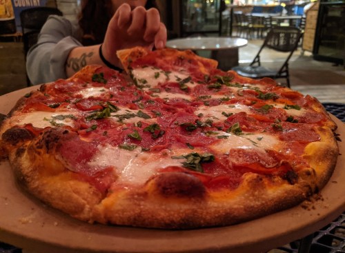 A hand reaches for a slice of pepperoni pizza topped with fresh herbs, served on a wooden platter.