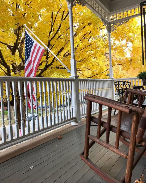 A porch with a wooden rocking chair and an American flag, surrounded by vibrant yellow autumn leaves.