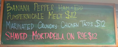 Chalkboard menu featuring three sandwich options: Ham & Egg, Chicken Tacos, and Mortadella on Rye, each $12.