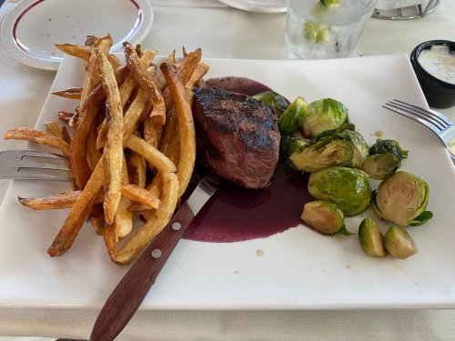 A plate with grilled steak, crispy fries, and Brussels sprouts, accompanied by a small bowl of sauce.