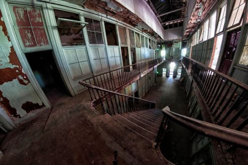 Abandoned interior of a building with peeling paint, a staircase, and large windows letting in greenish light.