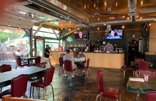 Interior of a casual restaurant with tables, a bar area, and a TV showing a program, featuring patrons and warm lighting.