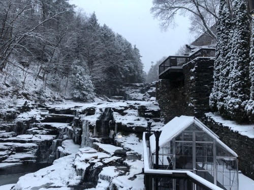 A snowy landscape featuring a waterfall, rocky terrain, and a glass greenhouse surrounded by trees.