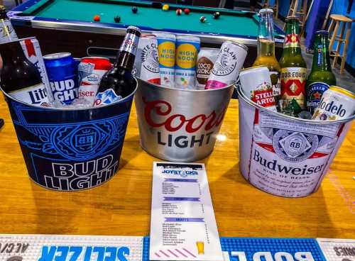 Three buckets filled with various beer bottles and cans, placed on a bar table with a pool table in the background.