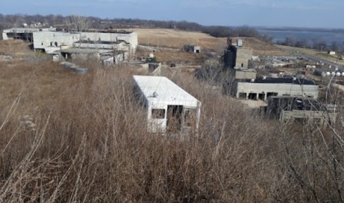 Abandoned buildings surrounded by overgrown grass and shrubs, with a river visible in the background under a clear sky.