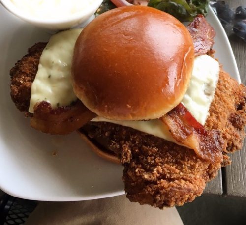 A crispy fried chicken sandwich with bacon and creamy sauce on a toasted bun, served with a side of coleslaw.
