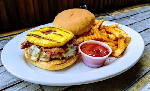A plate with a burger topped with bacon and grilled pineapple, served with crinkle fries and a side of ketchup.