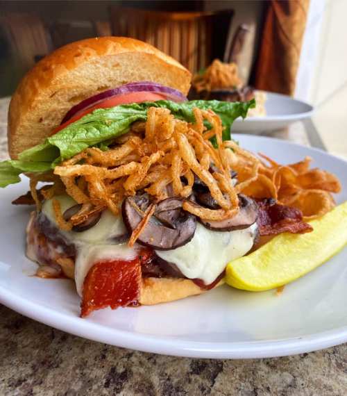 A delicious burger topped with lettuce, onions, mushrooms, cheese, and crispy onions, served with potato chips and a pickle.