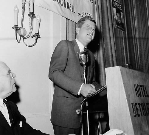 John F. Kennedy speaking at a podium with a microphone, while another man sits beside him at a hotel event.