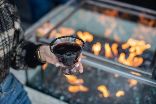 A hand holding a dark cocktail with a black rim, in front of a fire pit with flickering flames.