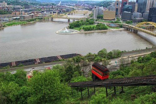 A red funicular railway descends a hillside overlooking a river and city skyline with bridges in the background.