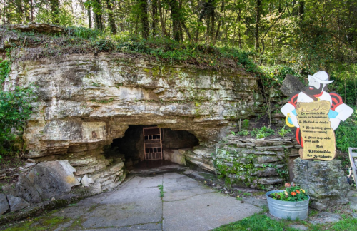 Entrance to a cave with a barred door, surrounded by greenery and a colorful sign nearby.