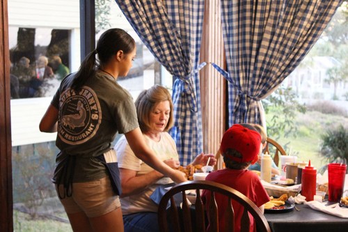 A waitress serves food to a woman and a child at a table by a window with checkered curtains.