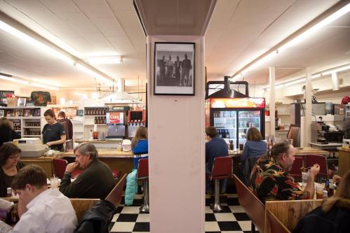 A busy diner with patrons at tables, a vintage photo on the wall, and staff serving in a cozy atmosphere.