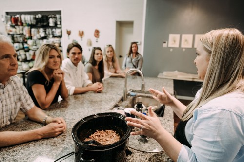 A group of people attentively listens to a woman speaking at a kitchen counter with a slow cooker.