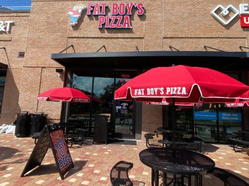 Exterior of Fat Boy's Pizza with red umbrellas over tables and a sign advertising pizza specials.