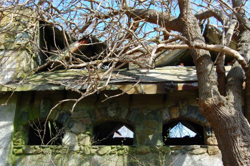 An abandoned stone building with a damaged roof, surrounded by bare branches against a clear blue sky.