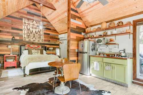 Cozy cabin interior with wooden walls, a bed, kitchen area, and a dining table, featuring rustic decor and warm lighting.